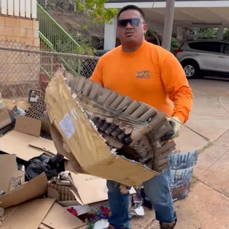 A crew member in an orange Unko's shirt carrying a broken panel away from a driveway pile of boxes and bags.